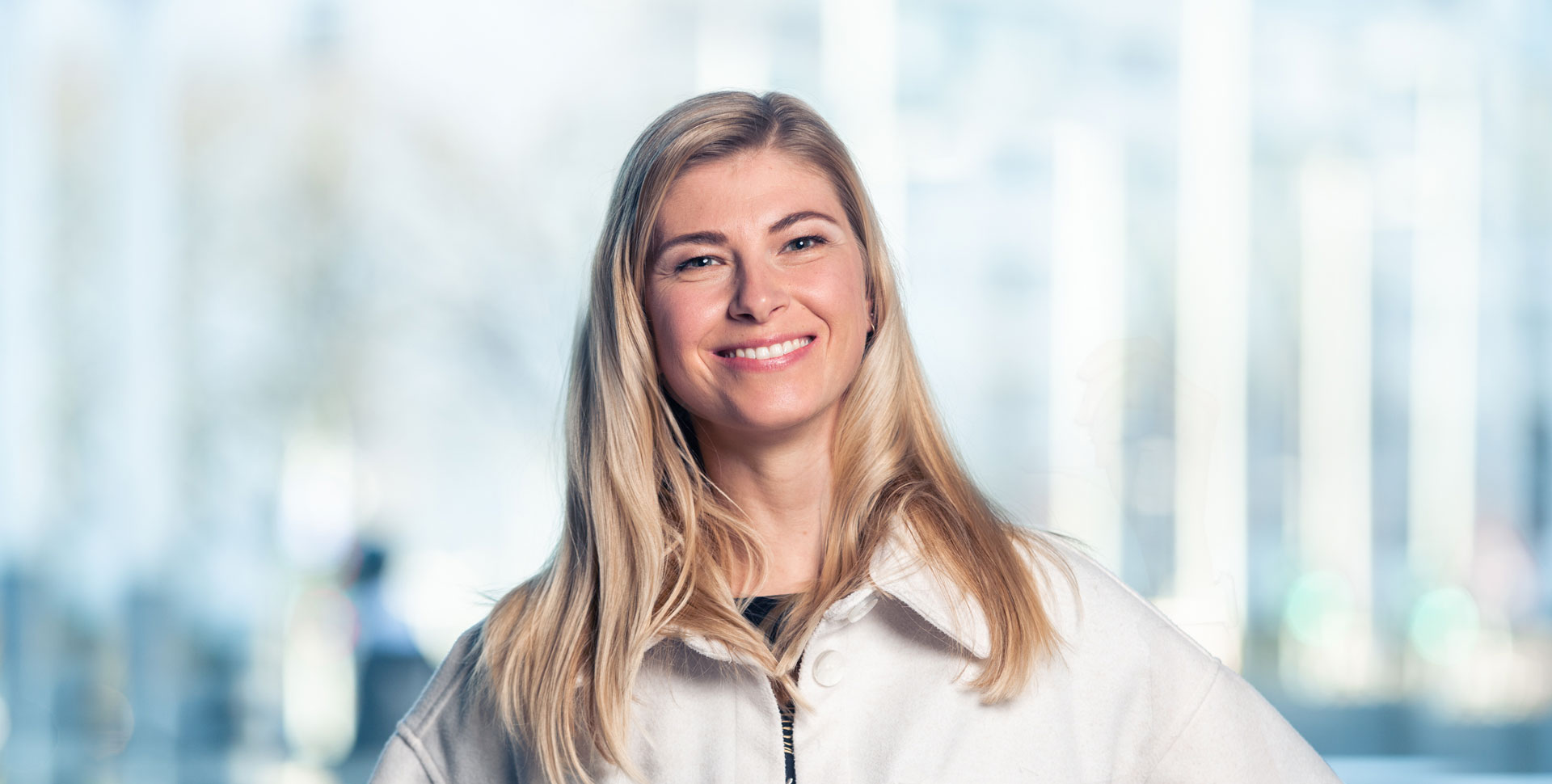 Lächelnde Frau mit langen blonden Haaren, die einen weißen Mantel trägt und in die Kamera schaut.