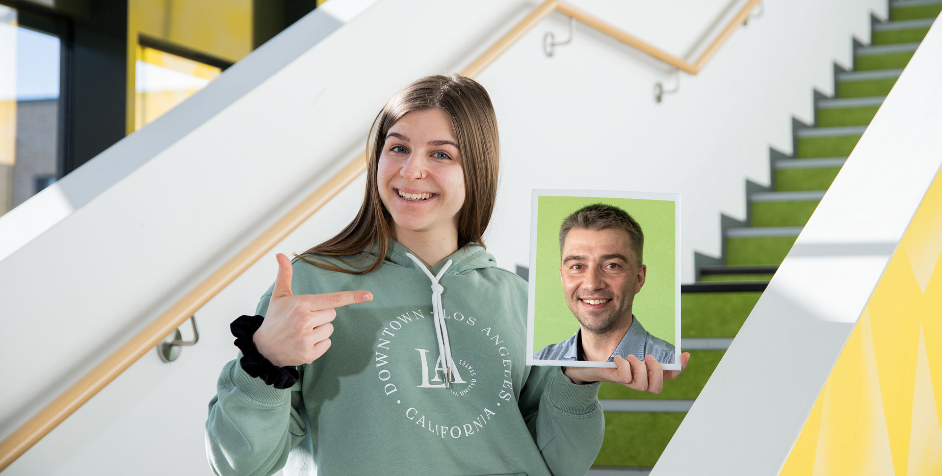 Junge Frau in einem grünen Hoodie, die auf ein Bild in einem Rahmen zeigt, während sie auf einer Treppe steht.