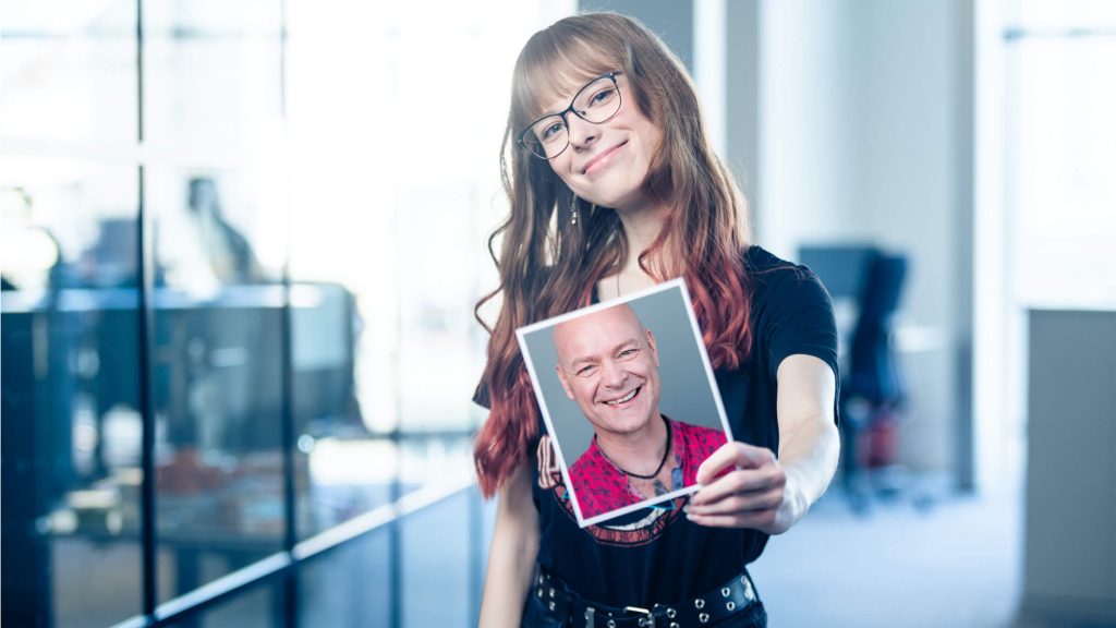 Frau hält ein Porträtfoto eines lächelnden Mannes in der Hand und steht in einem modernen Büro.