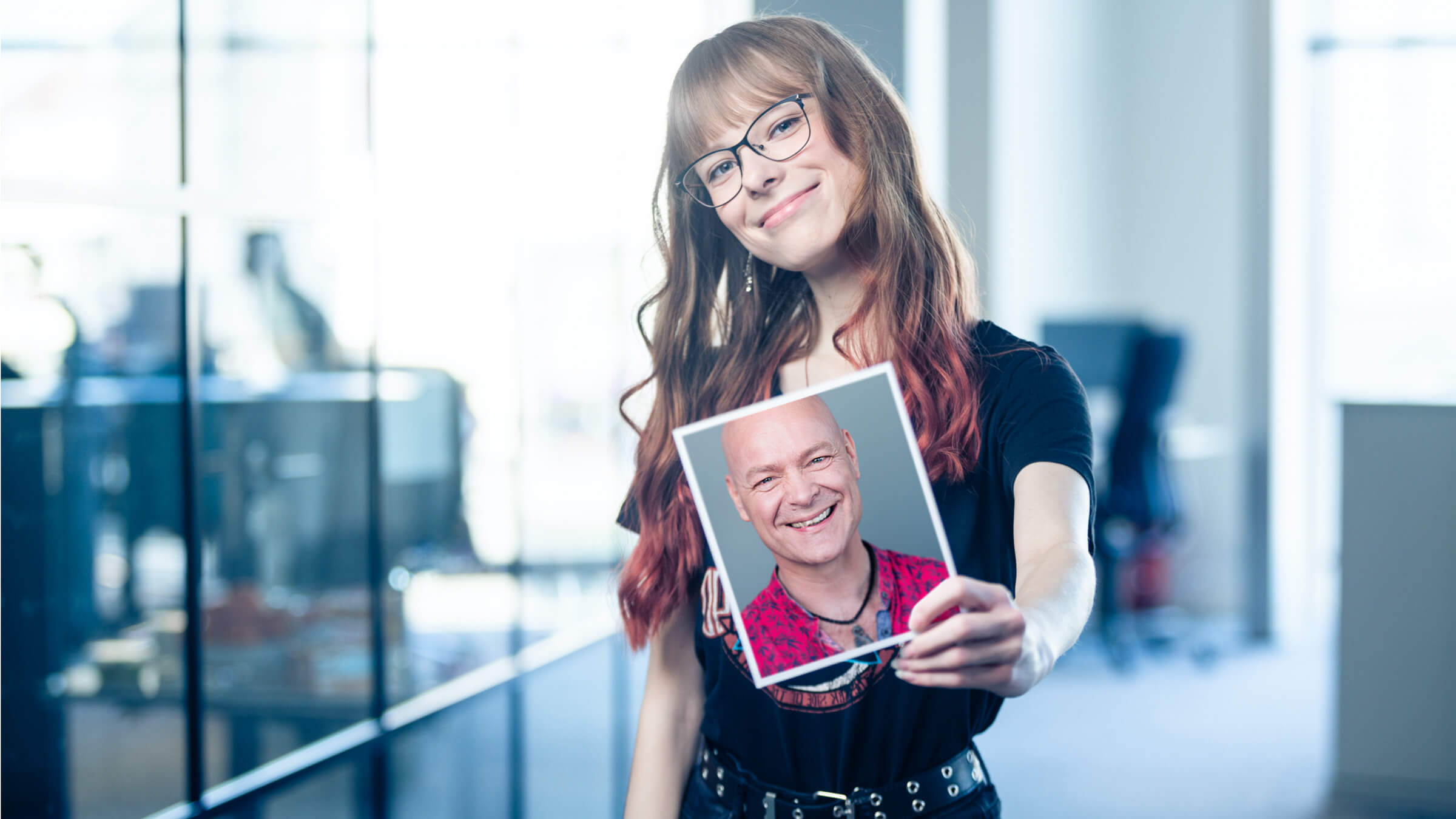 Frau hält ein Porträtfoto eines lächelnden Mannes in der Hand und steht in einem modernen Büro.