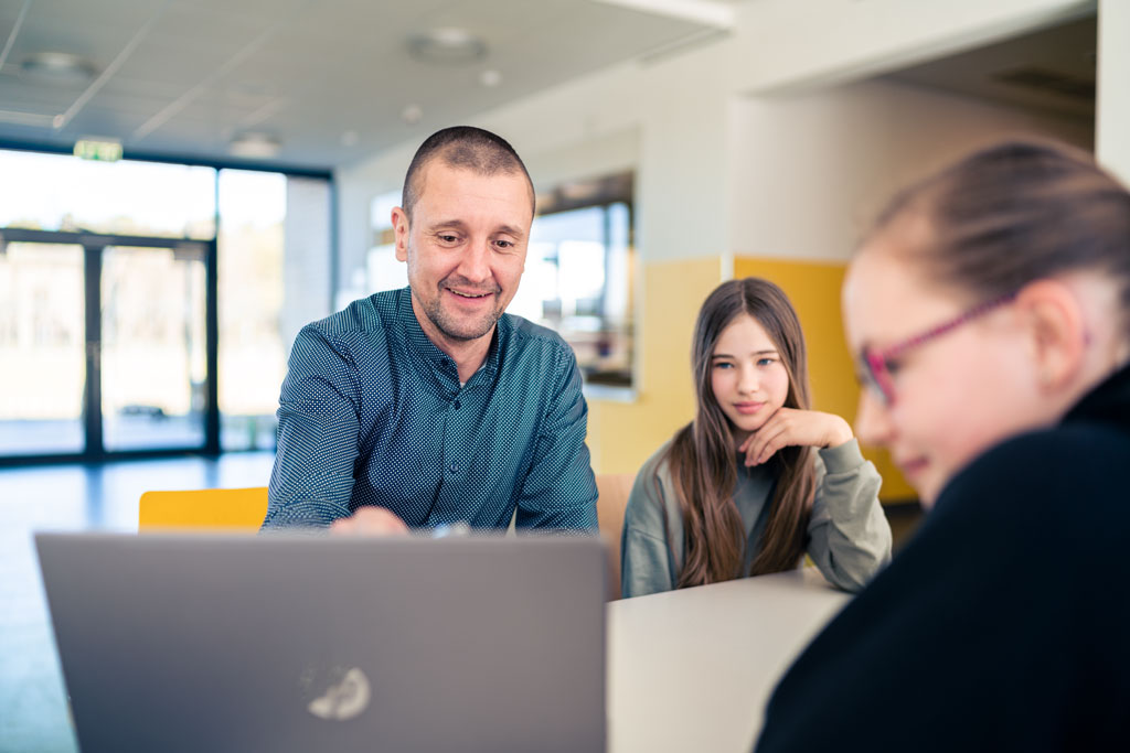 Junger Lehrer zeigt Schülerinnen am Tisch etwas auf dem Laptop
