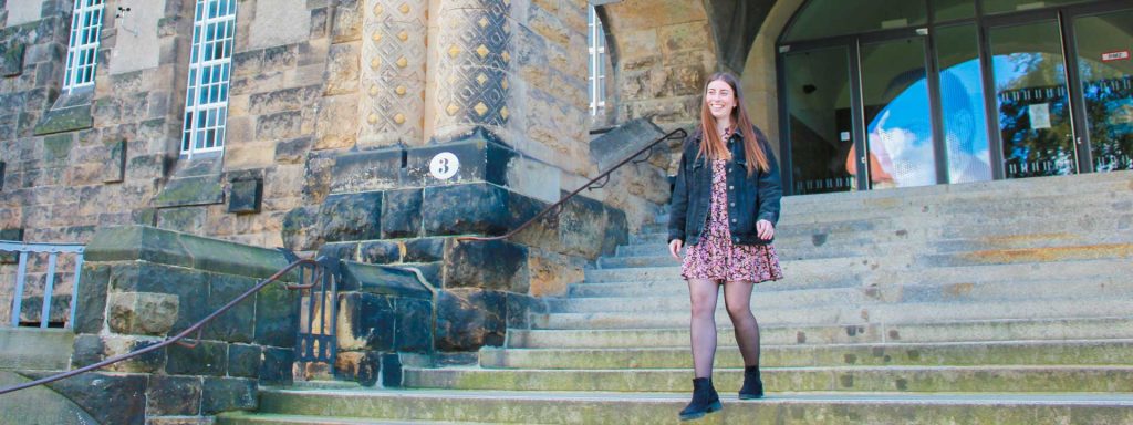 Junge Frau mit langen Haaren in einem Kleid, die die Stufen der Technischen Universität Dresden hinuntergeht.