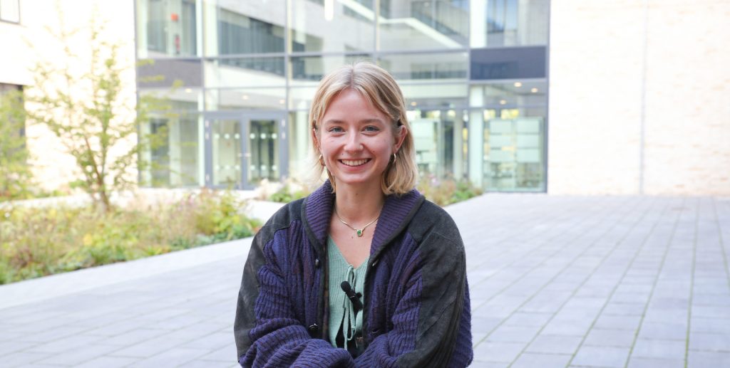Junge Frau mit blonden Haaren und einem lila Cardigan sitzt im Freien und lächelt in die Kamera.
