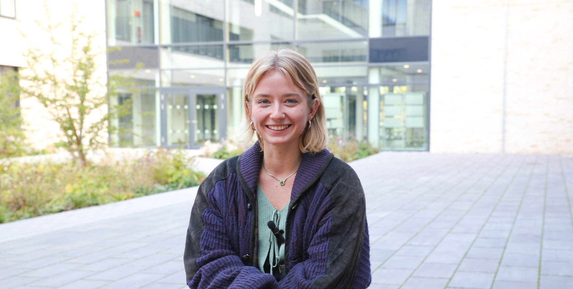 Junge Frau mit blonden Haaren und einem lila Cardigan sitzt im Freien und lächelt in die Kamera.