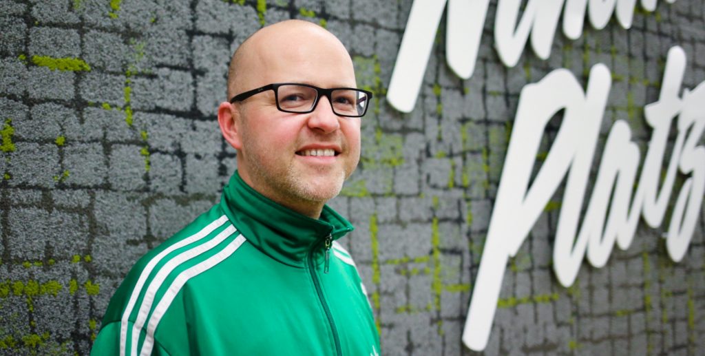 Mann mit Glatze und Brille trägt eine grüne Sportjacke mit weißen Streifen.