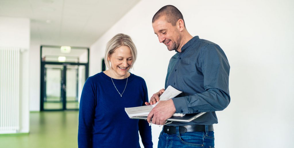 Zwei Lehrer stehen im Flur einer Schule und betrachten gemeinsam Unterlagen.