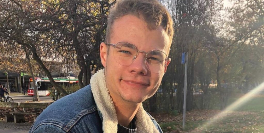 Junger Mann mit Brille und Jeansjacke, lächelnd in einem Park aufgenommen.