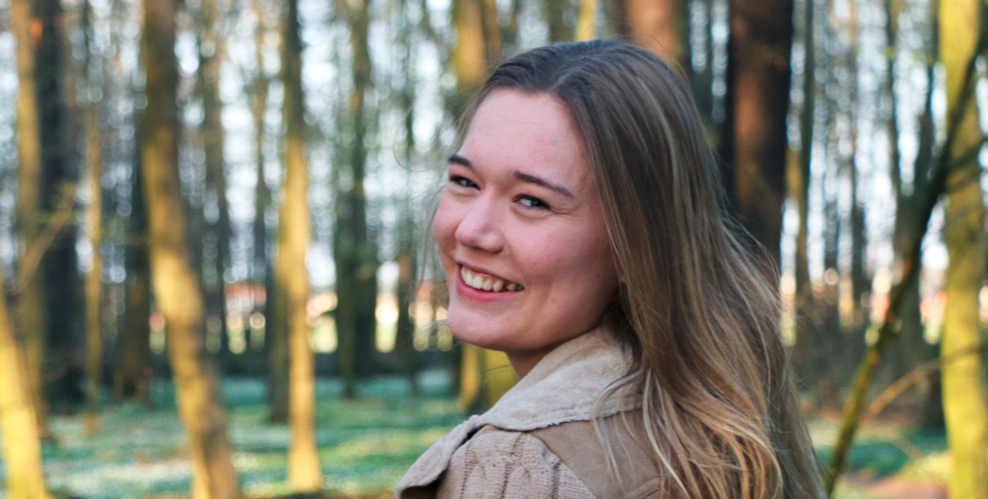 Junge Frau mit langen Haaren, die im Wald lächelt und in die Kamera schaut.