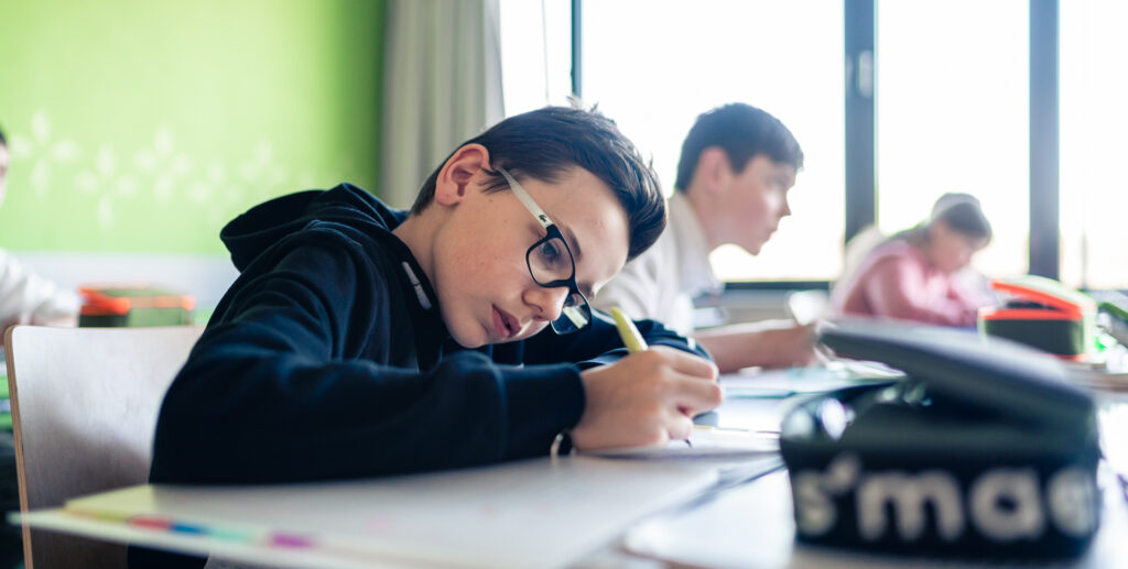 Junger Schüler mit Brille, der konzentriert in einem Heft schreibt, während andere Schüler im Hintergrund arbeiten.