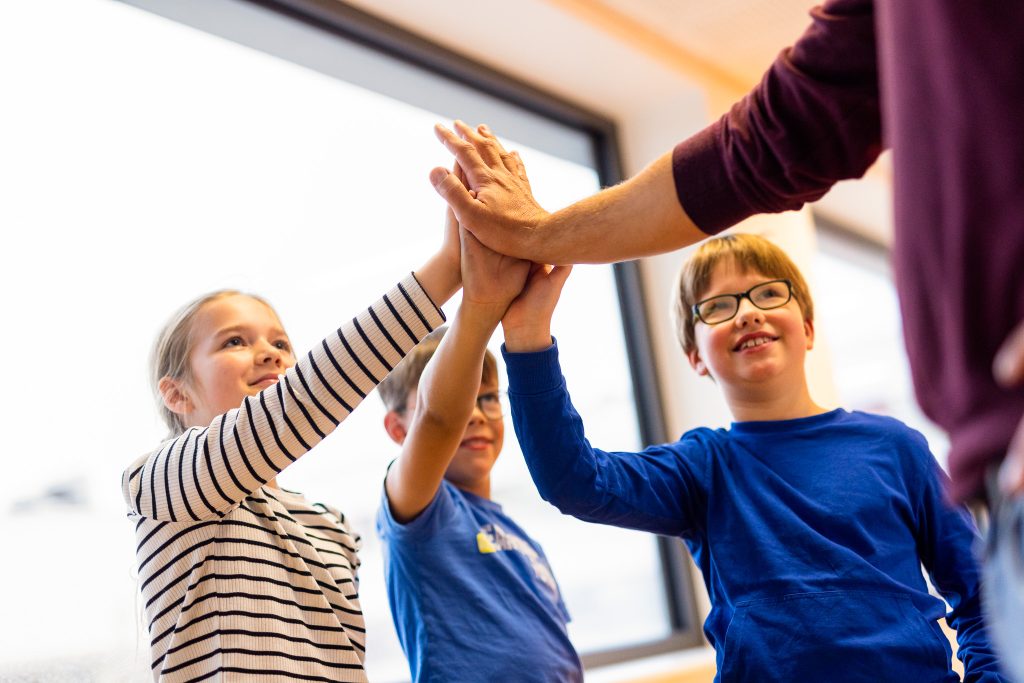 Drei Kinder geben sich im Klassenzimmer ein High-Five, während ein Erwachsener mit einer Hand beteiligt ist.