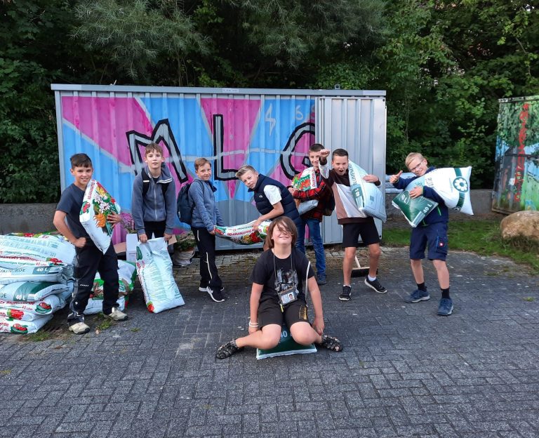 Eine Gruppe von Kindern hält Säcke mit Blumenerde vor einer bunten Wand.