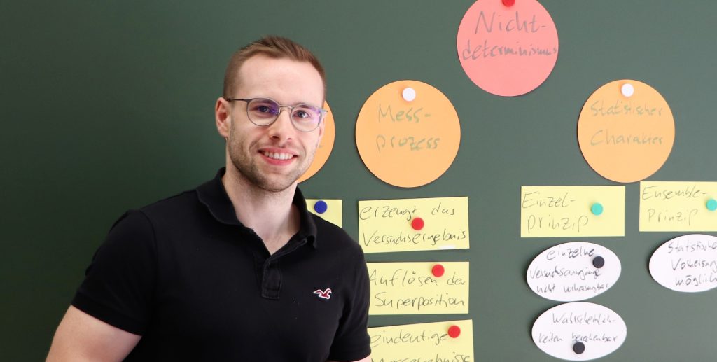 Mann mit Brille steht vor einer grünen Tafel mit bunten Notizen und Texten in einem Bildungsumfeld.