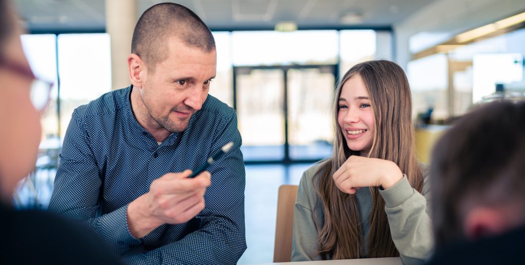 Ein Lehrer spricht mit einer Schülerin, während andere Schüler im Hintergrund sitzen.