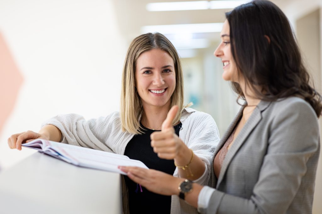 Zwei junge Lehrerinnen stehen in der Schulhalle, die eine schaut direkt in die Kamera, hebt den Daumen