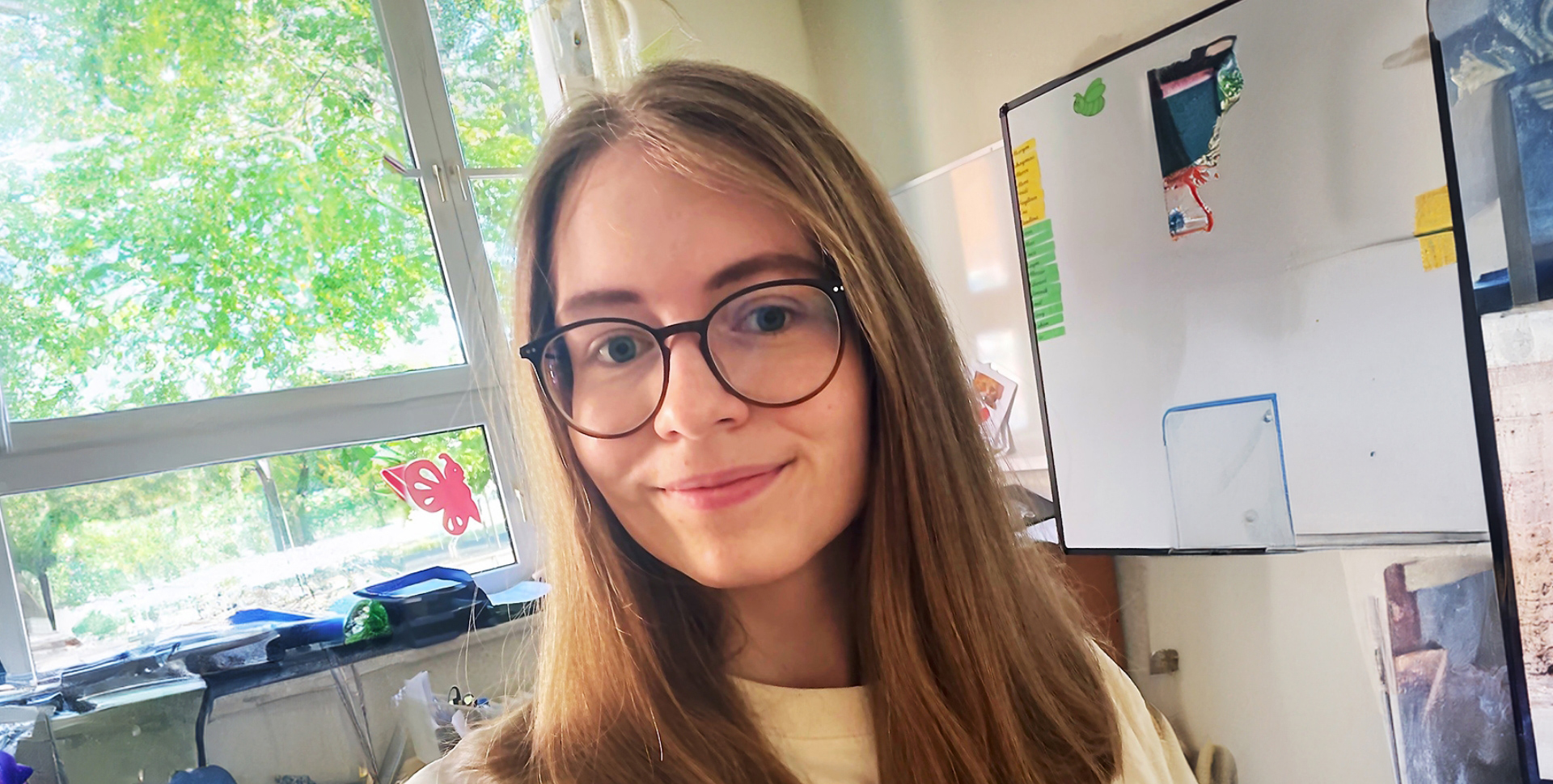 Junge Frau mit langen Haaren und Brille, die in einem Raum mit Fenstern steht.