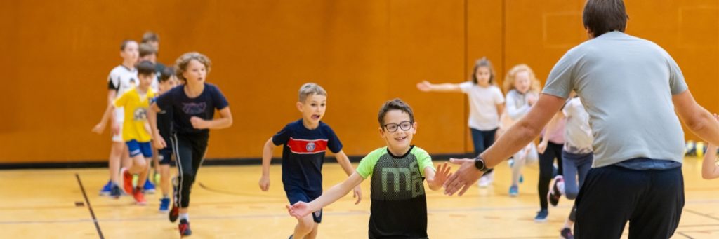 Schülerinnen und Schüler laufen in zwei Reihen auf ihren Sportlehrer bei einem Abklatschspiel zu. Ein Junge berührt beinah die Hand des Lehrers, der im Bild mit seiner Rückenansicht zu sehen ist.