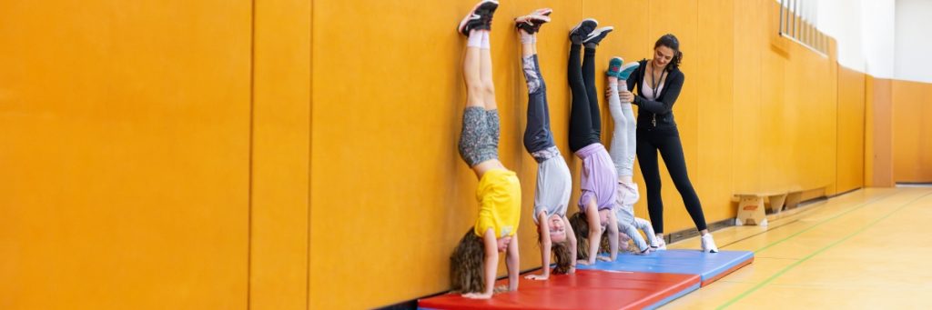 Vier junge Schülerinnen machen einen Handstand an einer Turnhallenwand. Die Lehrerin steht neben der rechten von ihnen und gibt ihr Hilfestellung.