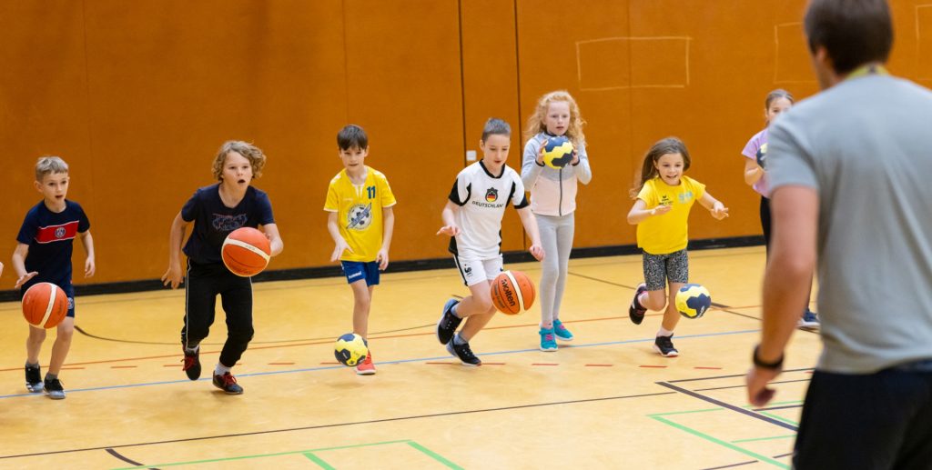 Eine Reihe von jungen Schülerinnen und Schülern läuft mit Basketbällen in einer Turnhalle auf den Sportlehrer zu, der rechts im Bild in der Rückenansicht zu sehen ist.