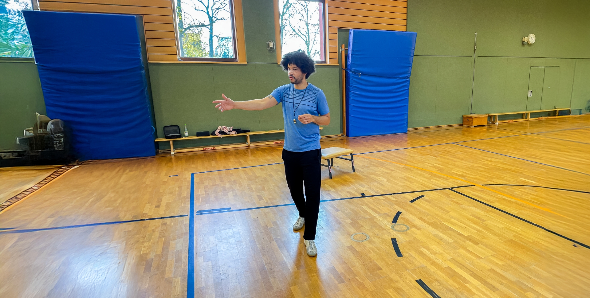 Sportlehrer Steve Hoppe weist in einer Turnhalle mit seinem rechten Arm auf eine Person oder Aktion, die im Bild aber nicht zu sehen ist.