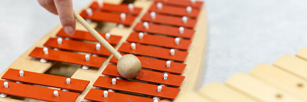 Hand schlägt mit einem Schlägel auf die roten Klangplatten eines Xylophons.