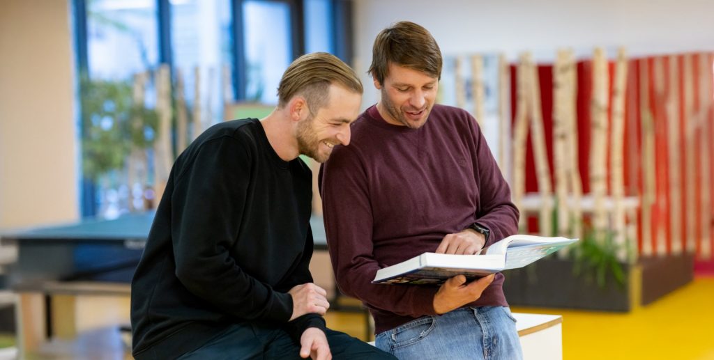 Zwei Männer sitzen nebeneinander und betrachten ein aufgeschlagenes Buch in einem Innenraum einer Schule.
