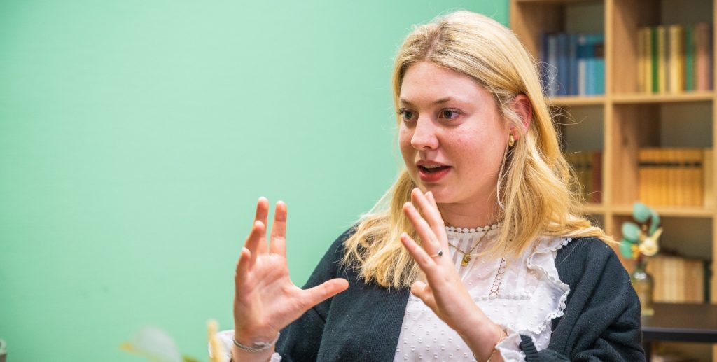 Junge Frau mit blonden Haaren und einem schwarzen Oberteil, die gestikulierend spricht.