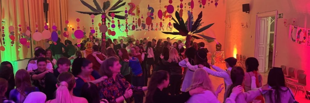 Schulball an der Petrischule in Leipzig: Schülerinnen und Schüler und andere Gäste des Balls tanzen eine Polonaise. Der Raum ist in Gelb, Orange, Rot und Pink erleuchtet und im Hintergrund sind Papp-Palmen zu sehen. Bunte Girlanden hängen von der Saaldecke herab.
