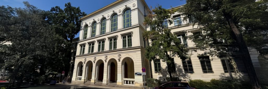 Die klassizistische Fassade der Petrischule in Leipzig ist zu sehen, umrahmt von Bäumen vor einem blauen Himmel.