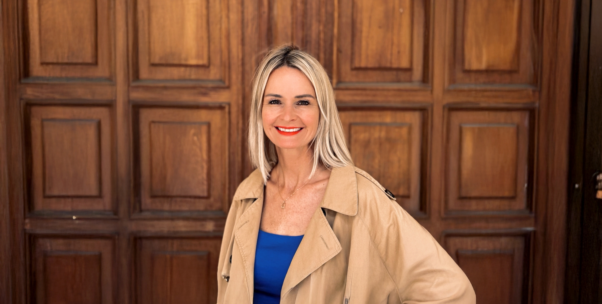 Eine blonde Frau, Marie-Christin Vollroth, steht im beiden Trench und blauen T-Shirt vor einer braunen Tür mit Holzkassetten.