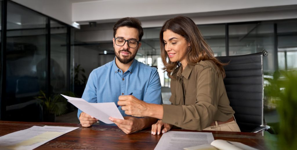 Zwei Personen, ein Mann und eine Frau, betrachten gemeinsam Dokumente am Tisch in einem modernen Büro.