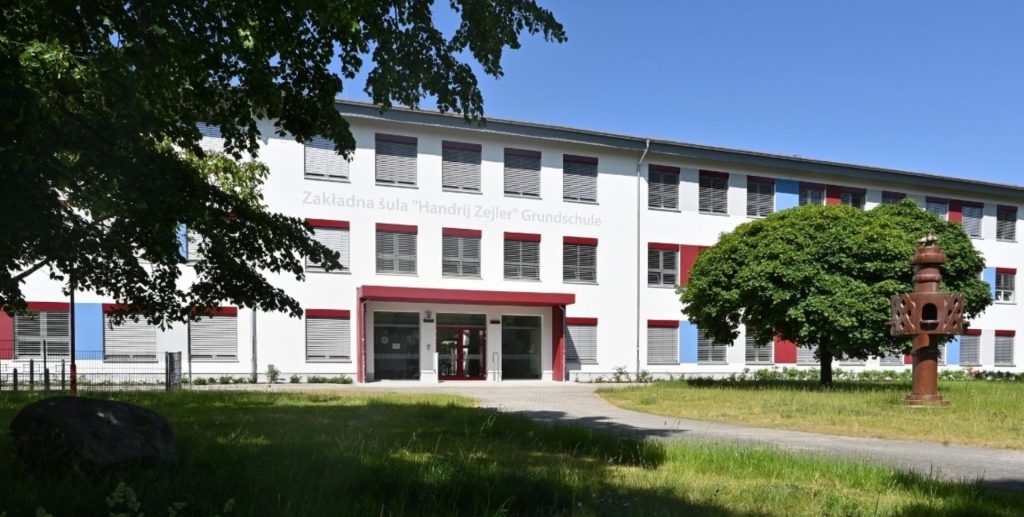 Es ist ein langgestrecktes weißes, zweistöckiges Gebäude vor blauem Himmel zu sehen. Über dem rot abgesetzten Eingansbereich steht auf der Fassade Zakladna Sula Handrij Zejler Grundschule.