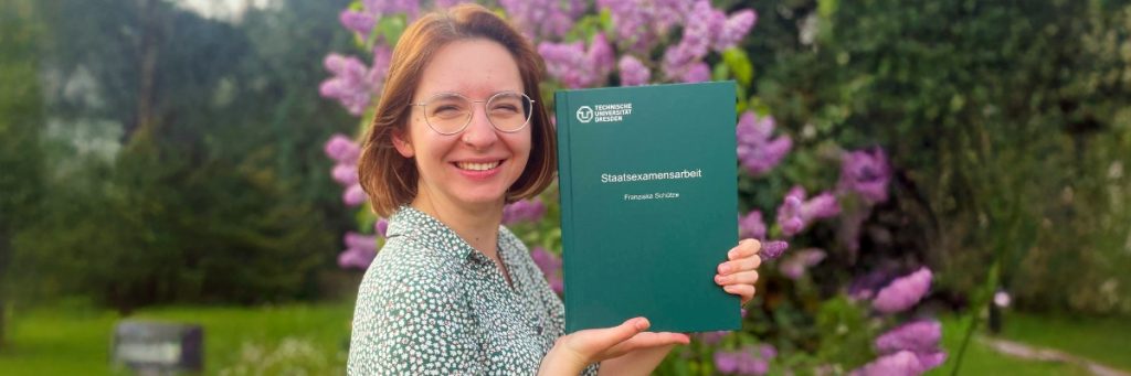 Franziska Schütze steht in einem Park vor einem lila Fliederbusch und hält ihre Staatsexamensarbeit der Technischen Universität in den Händen und präsentiert das Cover.