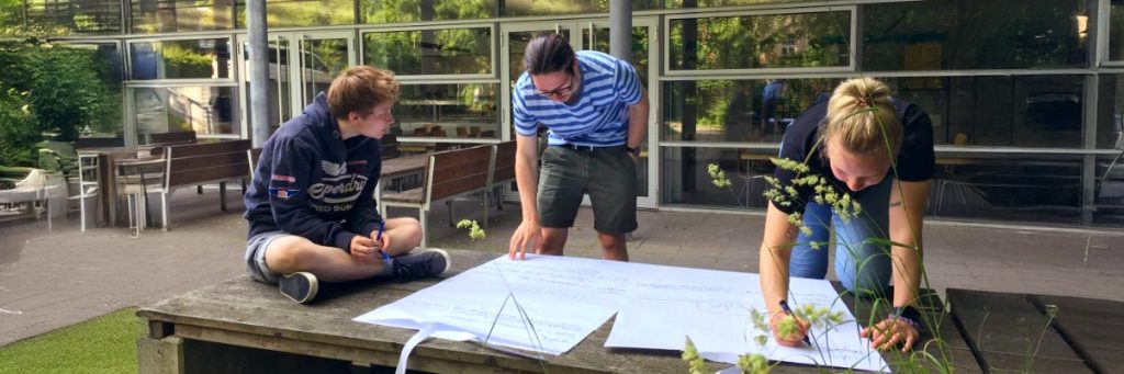 Zwei Männer und eine Frau beugen sich vor einer verglasten Wand über Flipchartbögen, die sie in Gruppenarbeit beschriften.