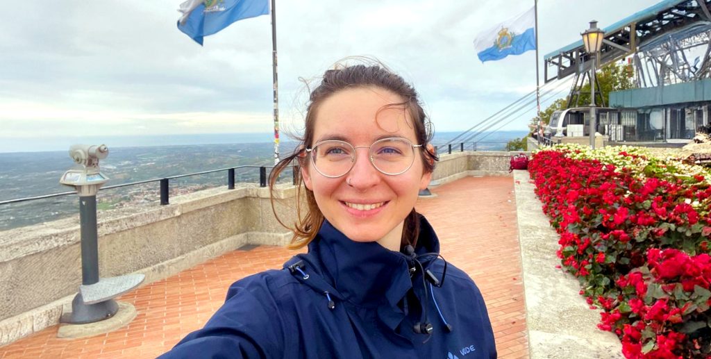 Eine Frau mit braunen, leicht verwehten Haaren steht an einer Strandpromenade. Rechts sind Rabatten von roten Bgeonien und anderen weißen Blumen zu sehen.