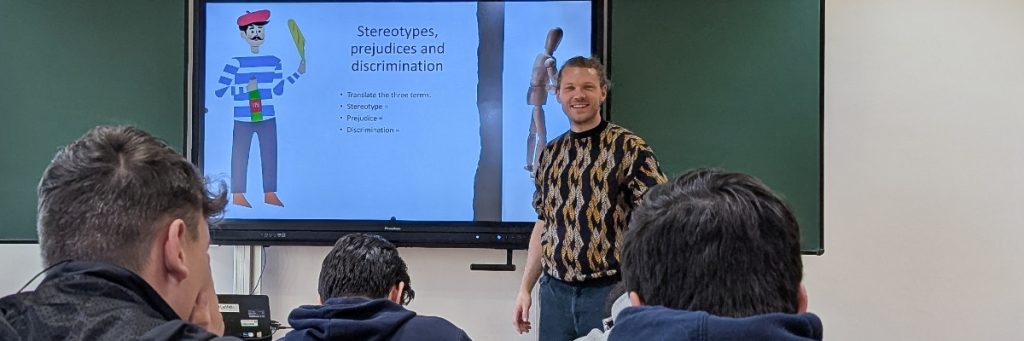 Lehrer Christian Kleinpeter steht vor einem Whiteboard auf dem ein Franzose mit Ringelshirt, roter Baskenmütze, Weinflasche und Baguette zu sehen ist. Daneben der Text: Stereotypes, prejudices and discrimination. Translate the three terms. Drei Schüler mit dunklen Haaren sind mit ihren Hinterköpfen im Vordergrund zu sehen.